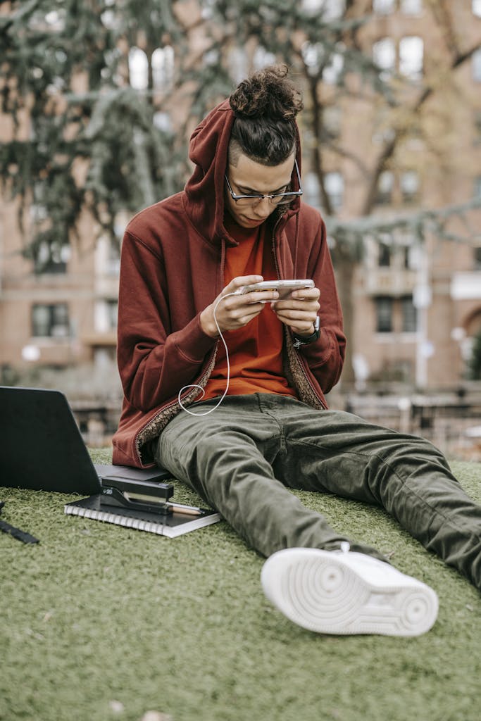 A young man in a hoodie sits outdoors, focused on his smartphone, with a laptop and notebook nearby.